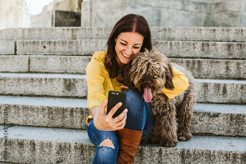 Young beautiful woman taking photograph of her sweet dog playfuly in a lovely park of the center of Madrid. Seated in stone stairs. Lifestyle photo