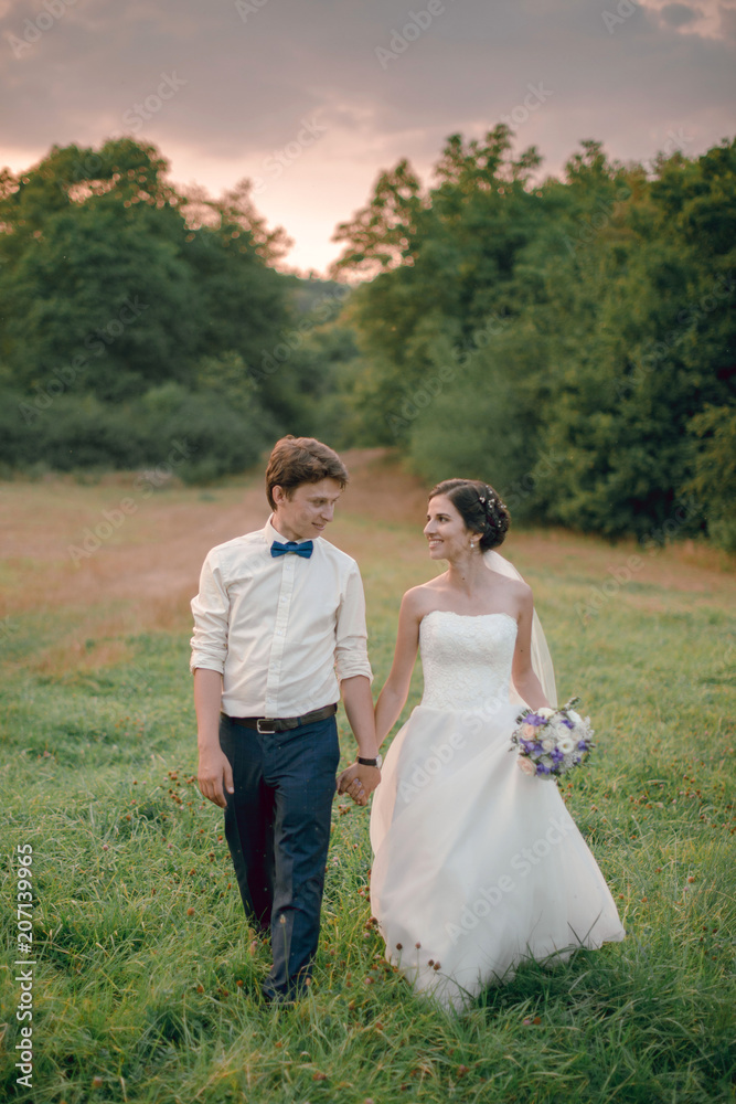 Happy young couple of groom and bride in white wedding dress with flower bouquet walking holding hands and smiling outdoors in the meadow in countryside