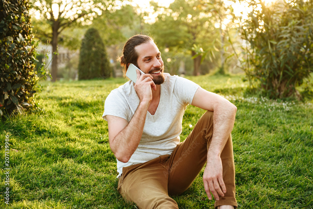 Picture of sociable young man in casual wear sitting on grass in green park, and speaking on smartphone with pleasure Picture of sociable young man in casual wear sitting on grass in green park, and speaking on smartphone with pleasure