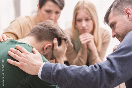 Fototapeta Naklejka Na Ścianę i Meble -  Close-up of a devastated young man holding his head in his hands and friends supporting him during group therapy