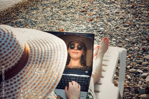 reflection in the laptop monitor of a young, smiling girl with glasses and hat, freelancer on the coast photo