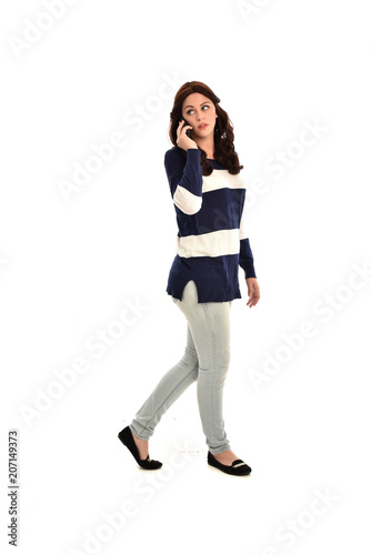 full length portrait of girl wearing striped blue and white jumper and jeans, holding a phone. standing pose on white studio background