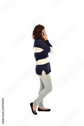 full length portrait of girl wearing striped blue and white jumper and jeans, holding a phone. standing pose on white studio background