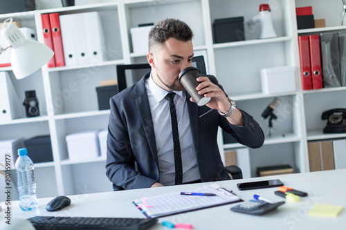 A man is sitting in the office at the table and drinking coffee from a glass.