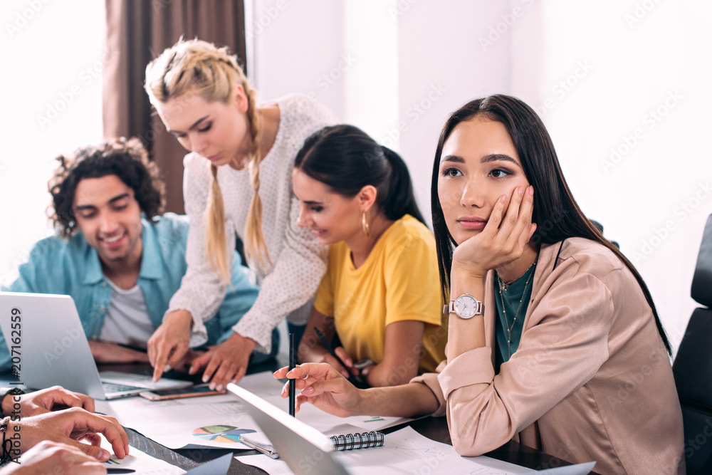 thoughtful asian businesswoman sitting at table while her colleagues having discussion behind at modern office thoughtful asian businesswoman sitting at table while her colleagues having discussion behind at modern office