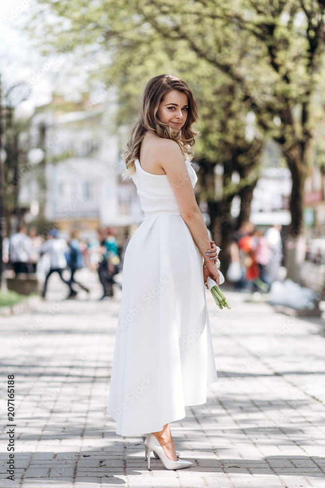 The charming bride keeps a wedding bouquet and stands on the street