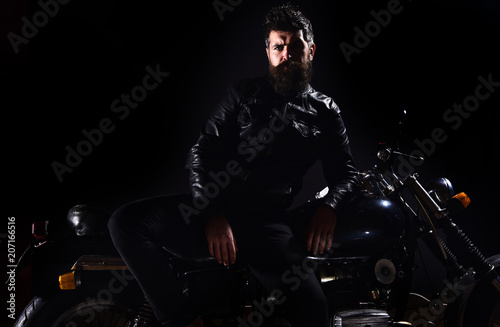 Macho, brutal biker in leather jacket stand near motorcycle at night time, copy space. Biker culture concept. Man with beard, biker in leather jacket lean on motor bike in darkness, black background.