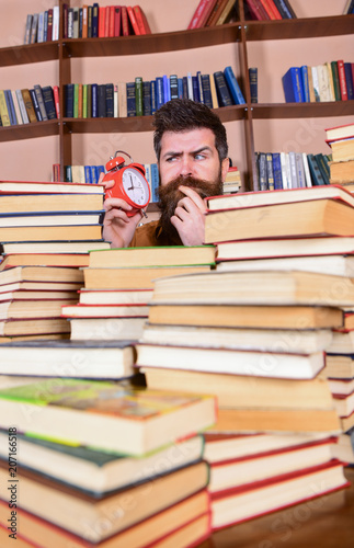 Teacher or student with beard studying in library. Man, scientist peeking out of piles of books with alarm clock. Man on thoughtful face looking at clock, bookshelves on background. Deadline concept.