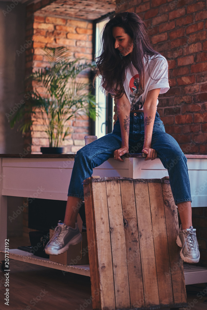 Cool modern girl wearing a white top and jeans sitting on a table in studio  with loft interior. Stock Photo | Adobe Stock