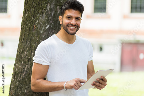 Young guy using a digital tablet lying against a tree in the park and enjoying the freedom of rhe wireless internet connection