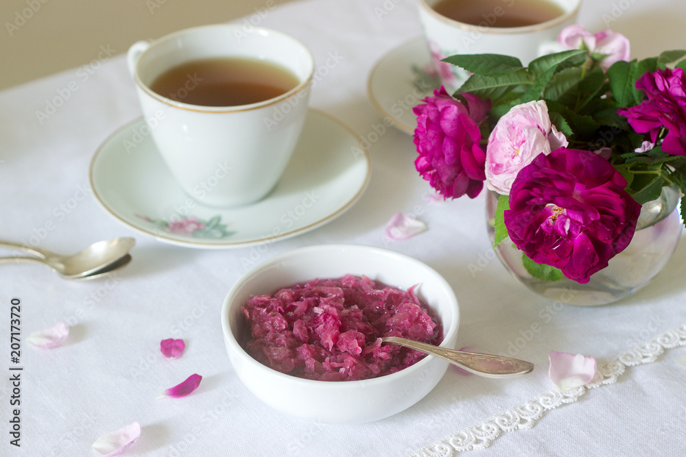 Jam from the petals of the Damascus rose, a cup of green tea and a vase of roses on a light table. Rustic style.