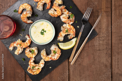 Overhead photo of plate of cooked shrimps on a dark rustic background, with a sauce and a glass of wine, with copy space