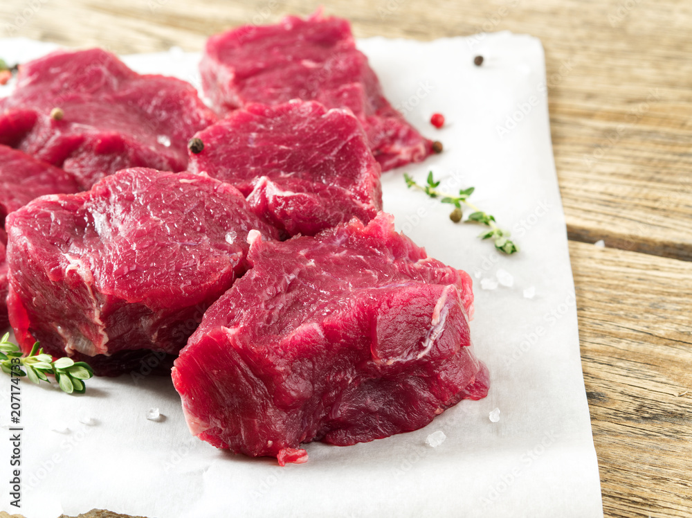 Pieces of raw meat. Raw beef with spices on white parchment paper on wooden rough rustic background, side view, close-up Pieces of raw meat. Raw beef with spices on white parchment paper on wooden rough rustic background, side view, close-up