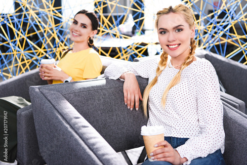 smiling businesswomen with paper cups of coffee at modern coworking office