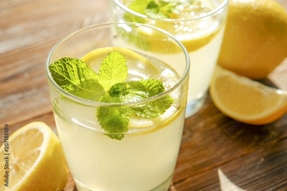 Two glasses full of ice cold refreshing lemonade beverage with mint leaves in sunlight on brown grunged wooden table, slices of ripe organic lemon, whole and halved. Background, copy space, close up. Two glasses full of ice cold refreshing lemonade beverage with mint leaves in sunlight on brown grunged wooden table, slices of ripe organic lemon, whole and halved. Background, copy space, close up.