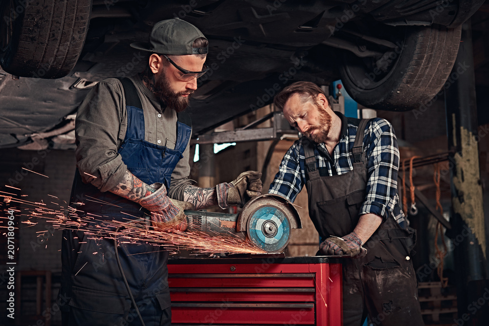 Two bearded auto mechanic in a uniform and safety glasses working with an angle grinder while standing under lifting car in repair garage. Two bearded auto mechanic in a uniform and safety glasses working with an angle grinder while standing under lifting car in repair garage.