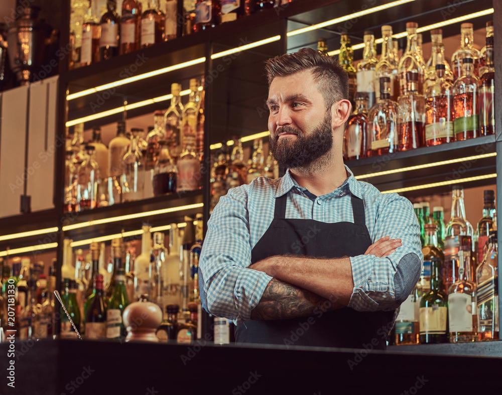 Stylish brutal bartender in a shirt and apron standing with crossed arms at bar counter background.