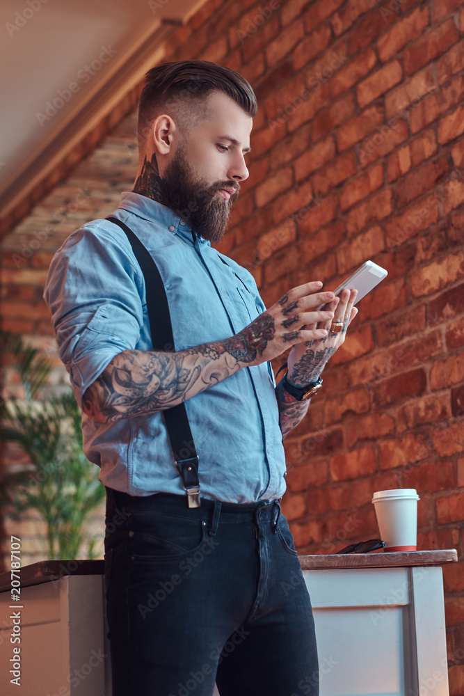 Old-fashioned tattooed hipster wearing a shirt and suspenders, in a  sunglasses, using a tablet while standing near a desk in an office with  loft interior. Stock Photo | Adobe Stock