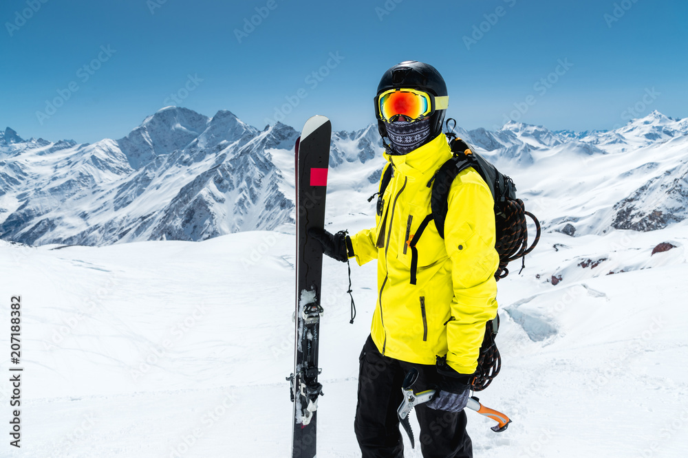 Close-up portrait of a skier wearing a protective helmet and glasses. A mask and scarf with an ice ax in his hands next to skis on the snow-capped mountains of the Caucasus. Skiing