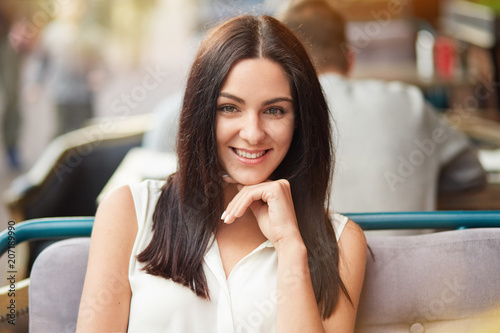 Portrait of happy brunette female with dark long hair, shining smile, keeps hand under chin, looks directly at camera. Attractive young woman recreats at cozy restaurant. People, leisure concept