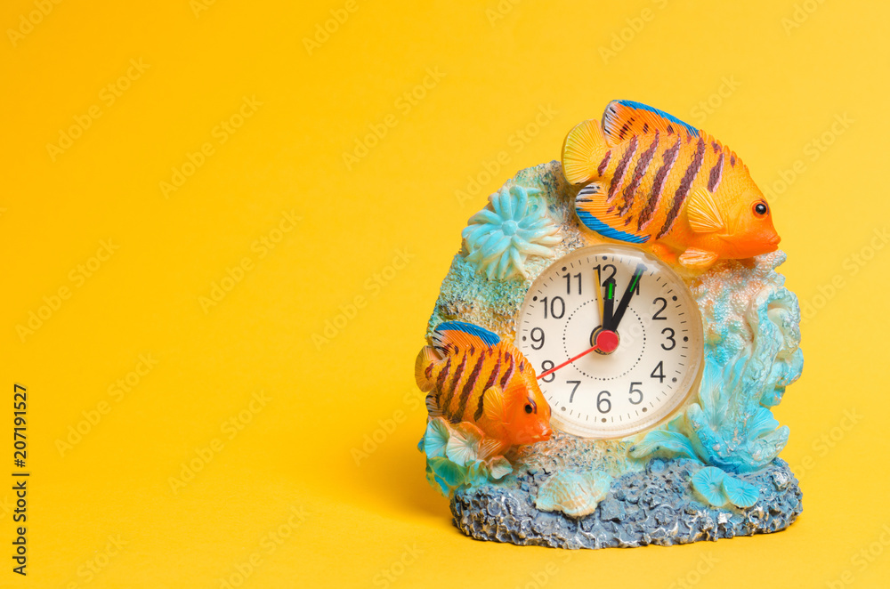 Fototapeta premium a clock with fish on a yellow background, a place for text. summer time. arrow points to the beginning of the first. alarm clock