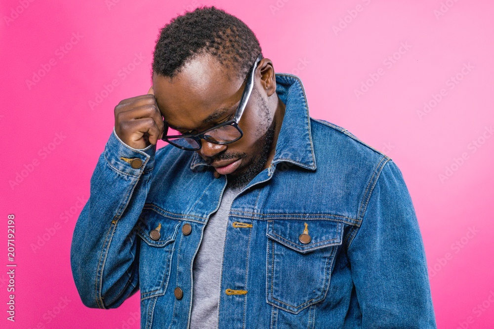 Thoughtful African man in glasses and jeans standing on a pink background brought his hand to his head Thoughtful African man in glasses and jeans standing on a pink background brought his hand to his head