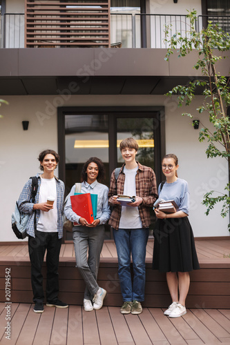 Young smiling students standing with books and folders in hands and happily looking in camera while spending time together in courtyard of university
