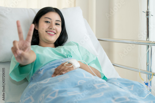 Young Asian patient woman lying at hospital bed with saline drip showing victory sign for cheerful. Drop of saline solution to help patient.