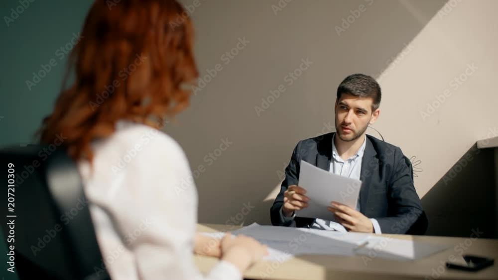 Young businessman talking with woman, holding documents sitting at table in company, entrepreneur communicating with female client during working day at desk in modern office. Concept: businesspeople Young businessman talking with woman, holding documents sitting at table in company, entrepreneur communicating with female client during working day at desk in modern office. Concept: businesspeople