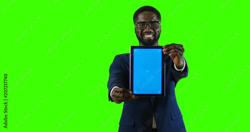 Portrait of the handsome smiled African American young businessman in the suit and tie posing and demonstrating a tablet computer in hands on the chroma key background. Green screen. Tracking motion Portrait of the handsome smiled African American young businessman in the suit and tie posing and demonstrating a tablet computer in hands on the chroma key background. Green screen. Tracking motion