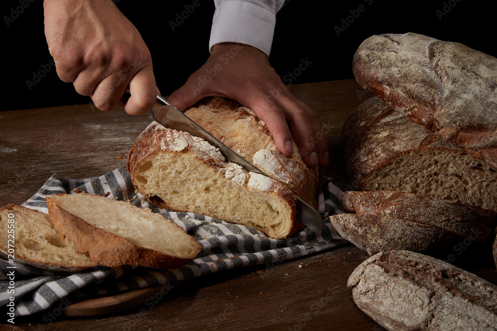 cropped shot of male baker cutting bread by knife on sackcloth on wooden table