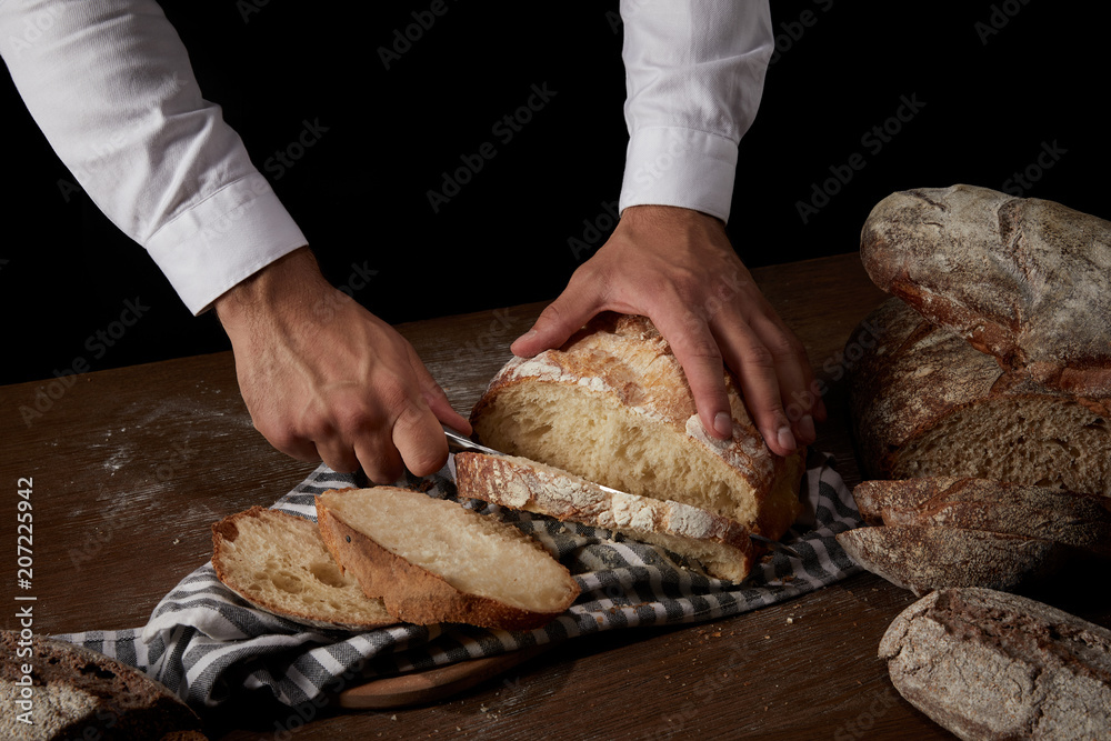 partial view of male baker cutting bread by knife on sackcloth on wooden table partial view of male baker cutting bread by knife on sackcloth on wooden table