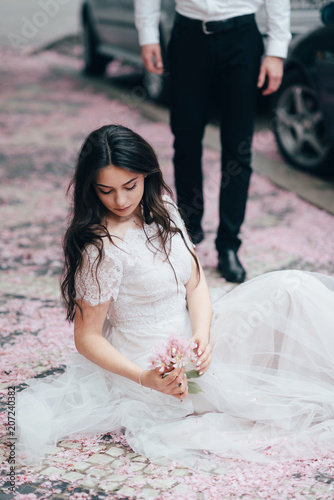 Portrait of a young beautiful brunette girl in white wedding dress sitting on a grass in a blooming spring garden. photo