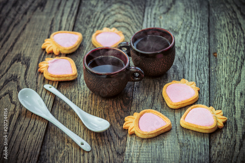 Ceramic cups with hot freshly brewed coffee, strawberry shape cookies with pink cream and white spoons on a wooden table. Sweet snack or breakfast.
