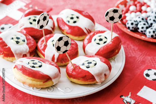 Doughnuts with red glaze and football decoration on red table cloth background. Chocolate candy football shaped on plate. Football toothpicks and flags with swiss cross. Football party in Switzerland.
