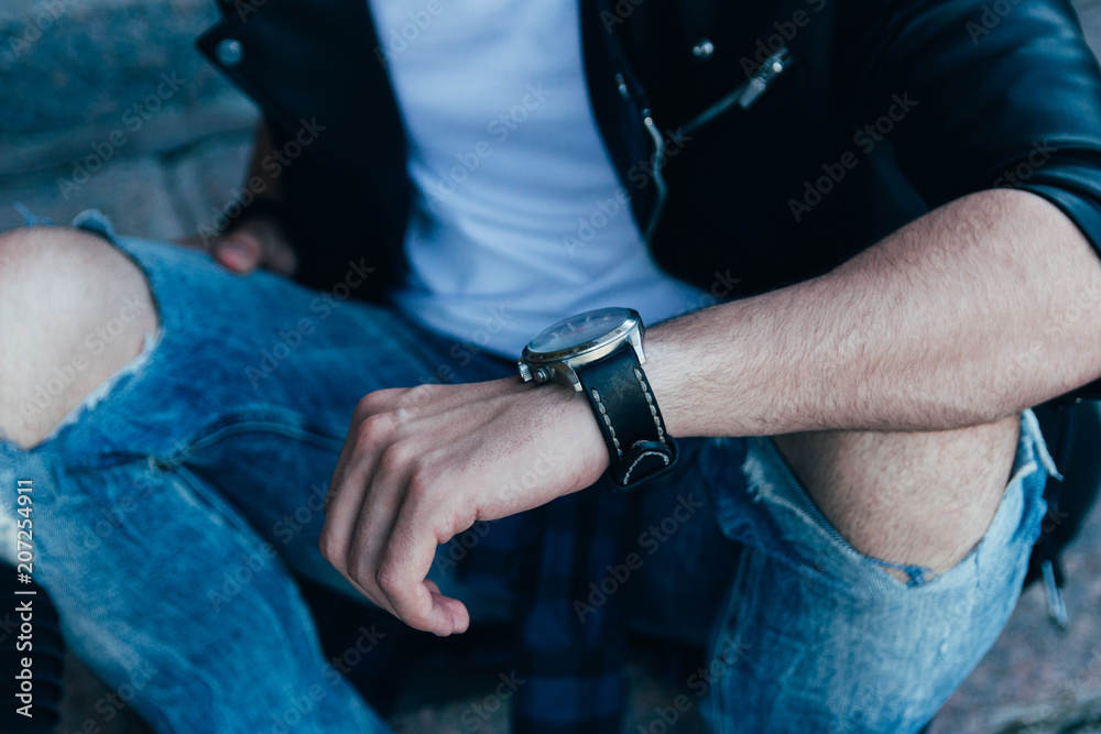 the man checks the time. in ragged jeans and a black jacket, street style. man's hand with a watch