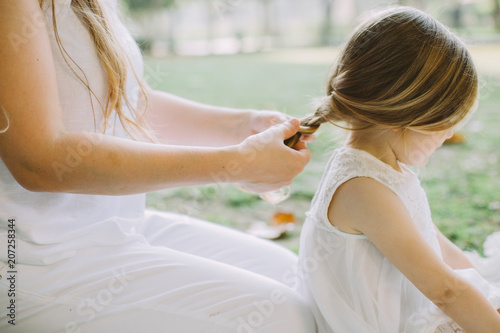 Portrait of beautiful young mother making braid to her adorable little daughter in park