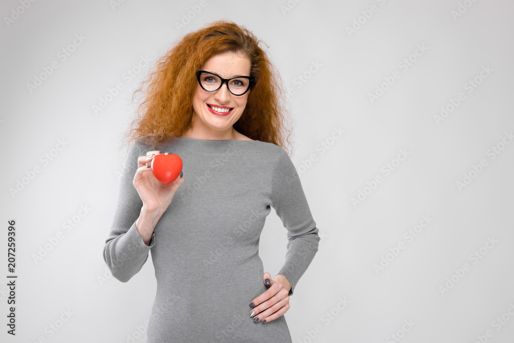 Charming young girl with red hair. A young girl in a gray sweater. Young girl on a gray background. A young girl with glasses is holding a red heart Charming young girl with red hair. A young girl in a gray sweater. Young girl on a gray background. A young girl with glasses is holding a red heart