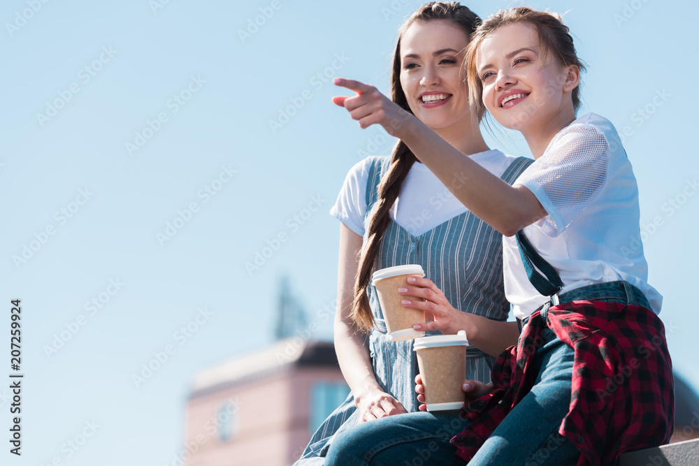 young woman pointing by finger to smiling female friend with paper cup of coffee young woman pointing by finger to smiling female friend with paper cup of coffee