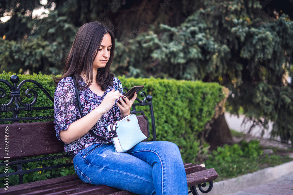 Girl with phone sitting on bench in park. Brunette woman with handbag