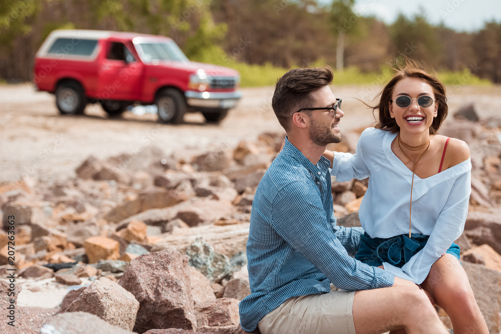 smiling couple sitting on rocks with red jeep on background