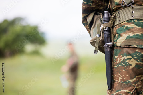 Soldier' body with camo clothing and knife in his belt. Close up behind view, blurred soldier and nature background, copyspace.