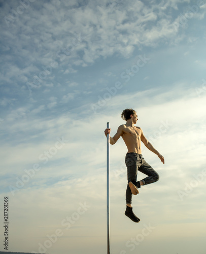 Athletic guy make acrobatic elements on pylon. Strong man dancer workout on pole. Young man dancing on pylon. Pole dance sport. Sexy macho man fly on blue sky background. In good shape.
