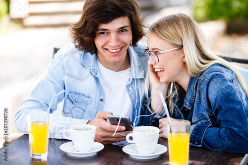 Young cheerful couple using headphones and smartphone for fun while sitting in cafe's garden