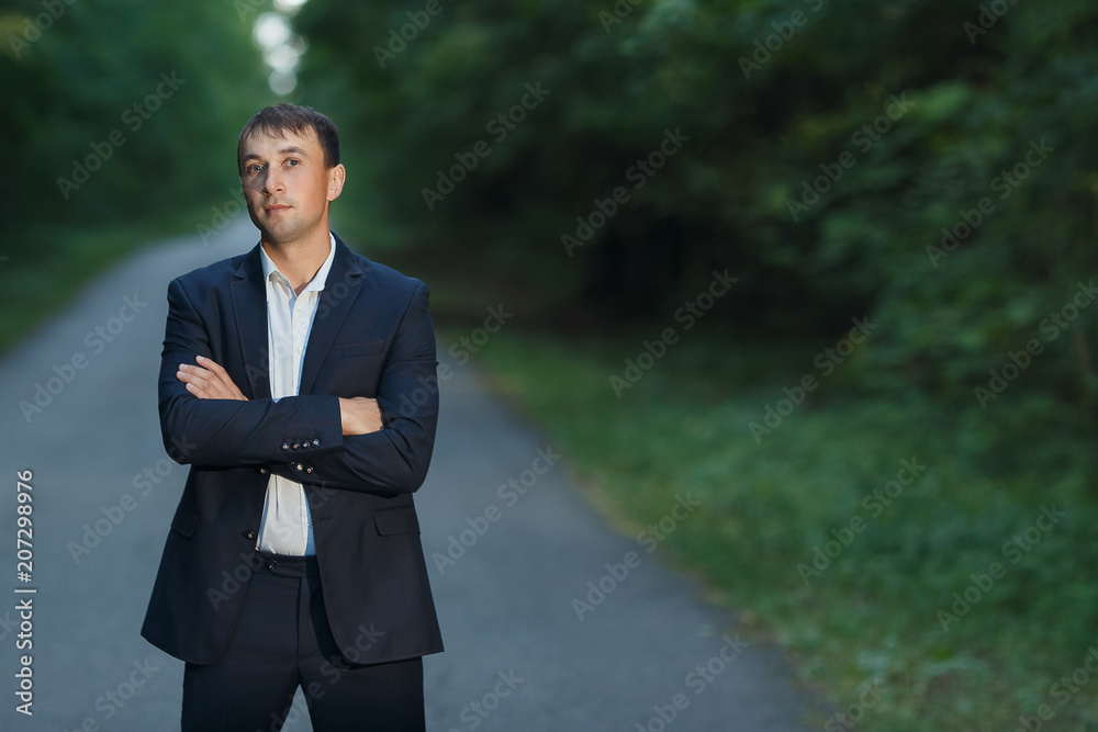 Young businessman in a classic suit with folded hands standing on the road in forest and looking at camera Young businessman in a classic suit with folded hands standing on the road in forest and looking at camera