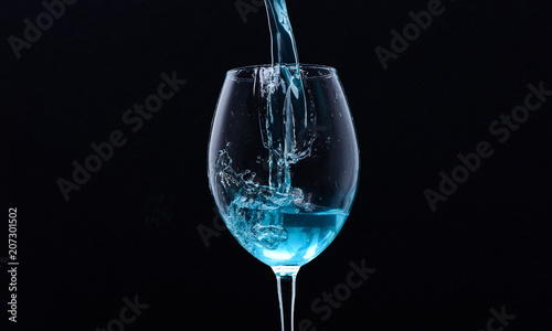 Wineglass filling with water with splashes on black background. Refreshing drink concept. Cocktail with blue liquid in glass. Glass with blue water pouring with liquid with splashes and drops