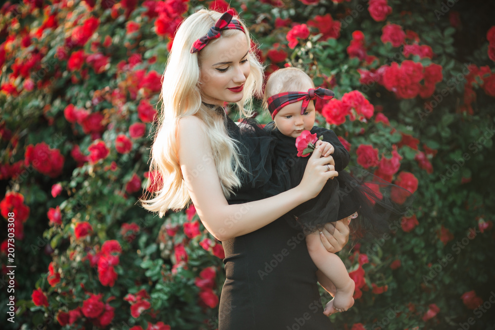Beautiful blonde woman with little baby girl in bush of red roses