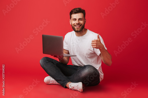 Photo of bearded caucasian guy in t-shirt and jeans sitting on floor with legs crossed and showing thumb up while holding laptop, isolated over red background photo
