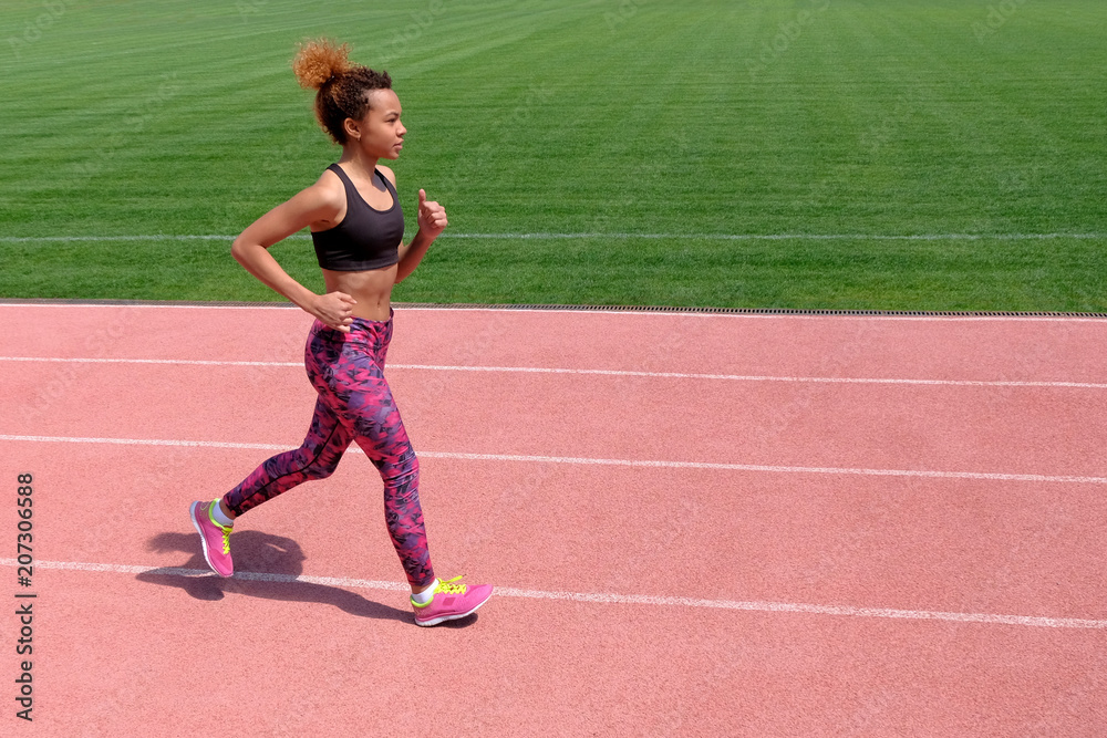 An African American in the summer sports. A young sexy sporty black girl runs along the pink path of the stadium next to the green grass.