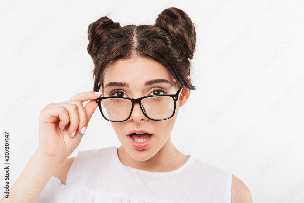 Photo closeup of surprised teenage girl 20s with double buns hairstyle and dental braces touching eyeglasses with curious look, isolated over white background